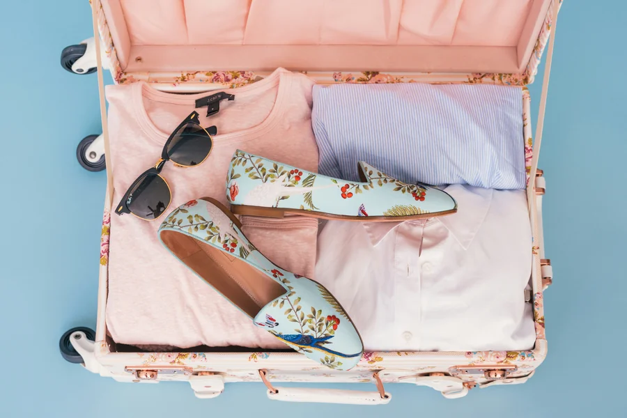 A pink floral suitcase with pink and light blue clothing inside, a pair of sunglasses, and floral summer shoes.